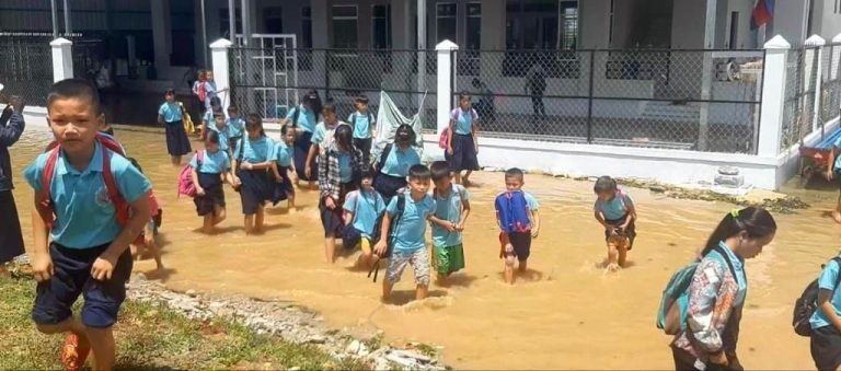 A group of Le Van Khanh students with backpacks wading through muddy floodwater outside the school
