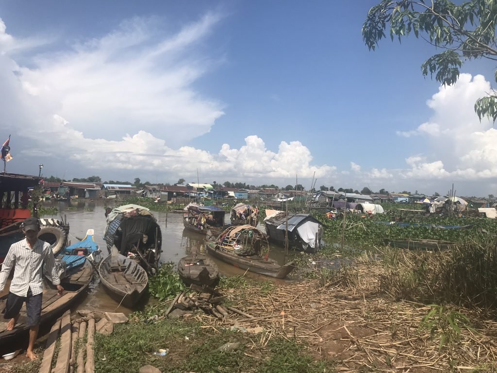 A panoramic view of the floating village on Tonle Sap Lake — small boats, makeshift shelters, and a man standing in the foreground