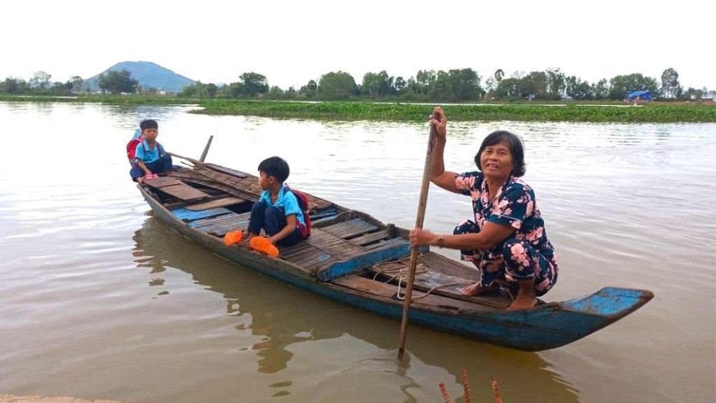 A woman paddling a wooden boat with two children in school uniforms, on the Tonle Sap River