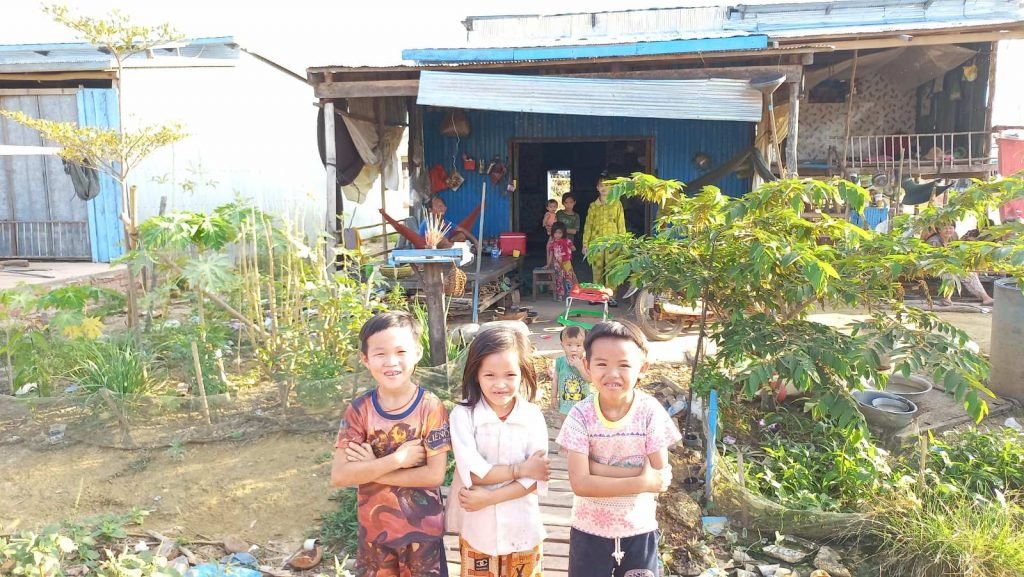 BeEm, My, and Nhi — three siblings — smiling outside their family home near the Tonle Sap River, with their younger siblings and parents in the background