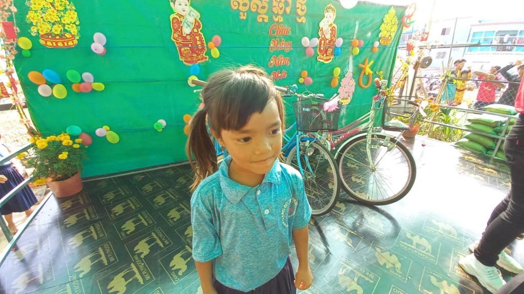 HoangMy, a young girl in a blue school uniform with her hair in a side ponytail, standing in front of her new bicycle against a Tết-decorated green wall