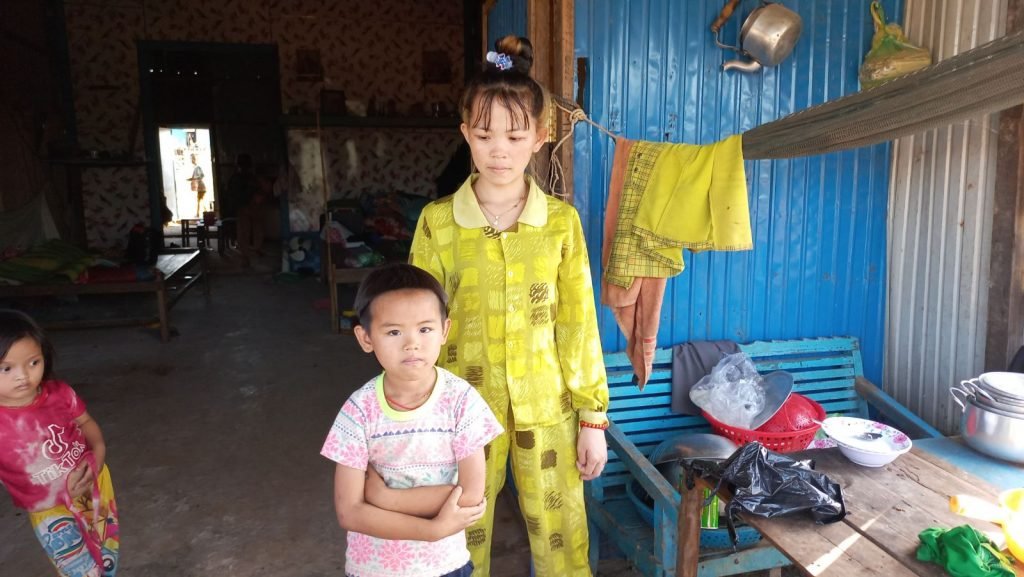 Inside a modest home in Tonle Sap — a young girl in yellow pajamas standing with her younger brother near the kitchen area