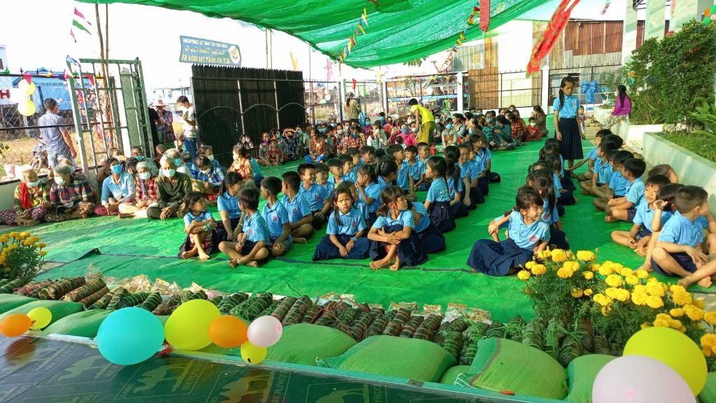 Students in blue uniforms sitting cross-legged on green tarp around bánh chưng and food gifts, waiting for the New Year celebration to begin