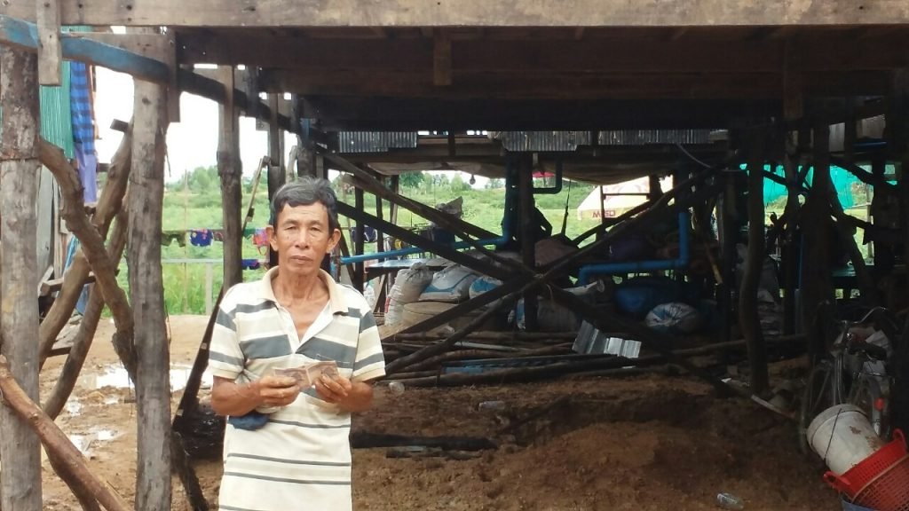 An older man standing under the elevated platform of his stilt house, where a new toilet has been installed thanks to IHM donor support