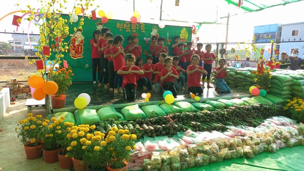 A group of children in matching red shirts kneeling in choreographed prayer pose during a Tết dance performance, with rice bags and gifts displayed in front of them