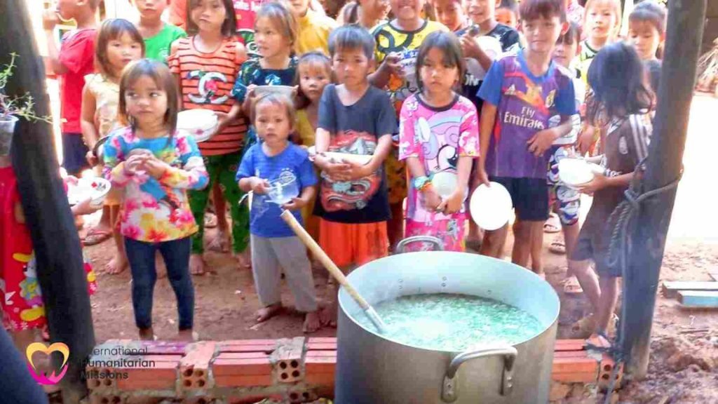 Children of Tonle Sap gathered with their bowls around a large pot of food at the IHM kitchen