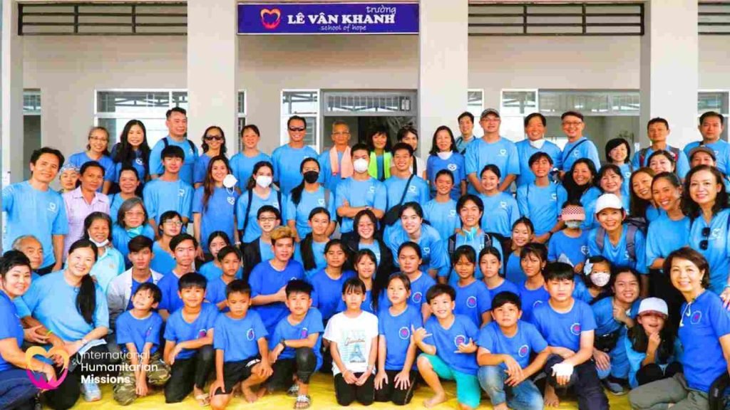 A large group photo of the IHM team together with Le Van Khanh students, all in matching blue shirts in front of the school sign